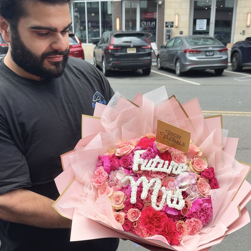 Man holding a bouquet of flowers with 'Future Mrs.' on it, street scene in the background