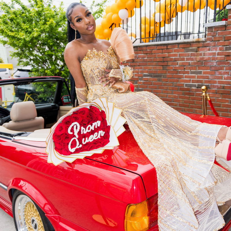 Woman in a gold dress sitting on a red car with 'Prom Queen' sign, surrounded by balloons.