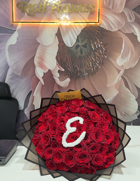 A large circular bouquet of red roses wrapped in black mesh, featuring a white flower letter 'E' in the center, displayed on a white counter in front of a floral wall mural and a glowing 'Rich Florals' sign.
